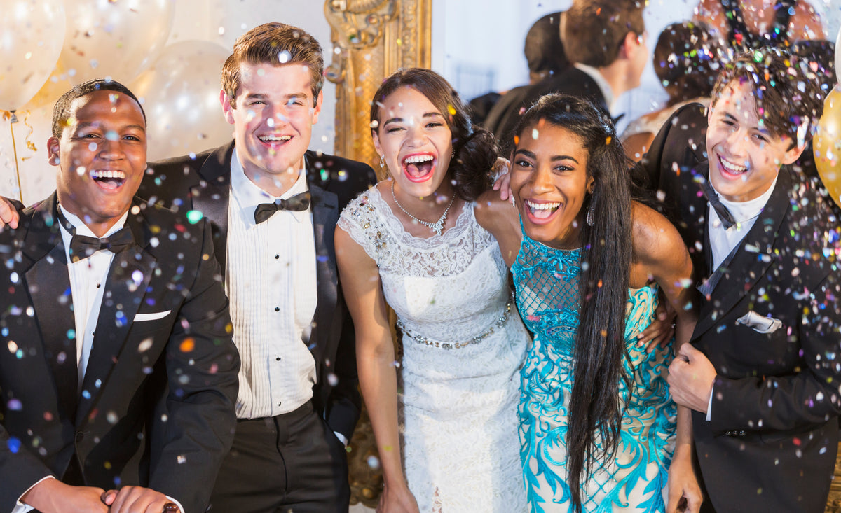 Group of teens in prom tuxedos and dresses laughing under confetti